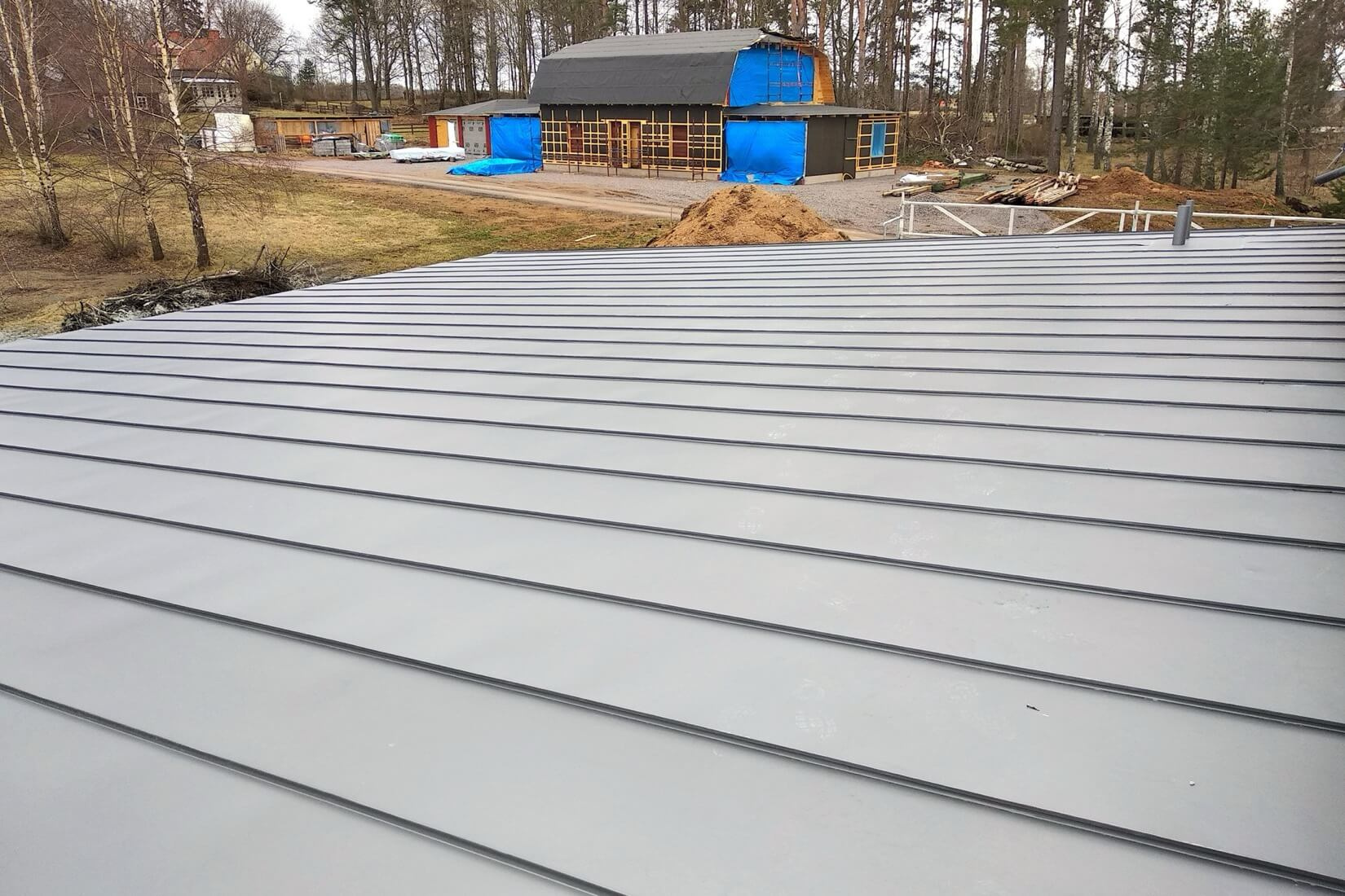 Vastikään asennettu harmaa metallikatto, jossa on kaistakate talon päällä, taustalla toinen rakenteilla oleva rakennus, joka osoittaa kattohanketta.